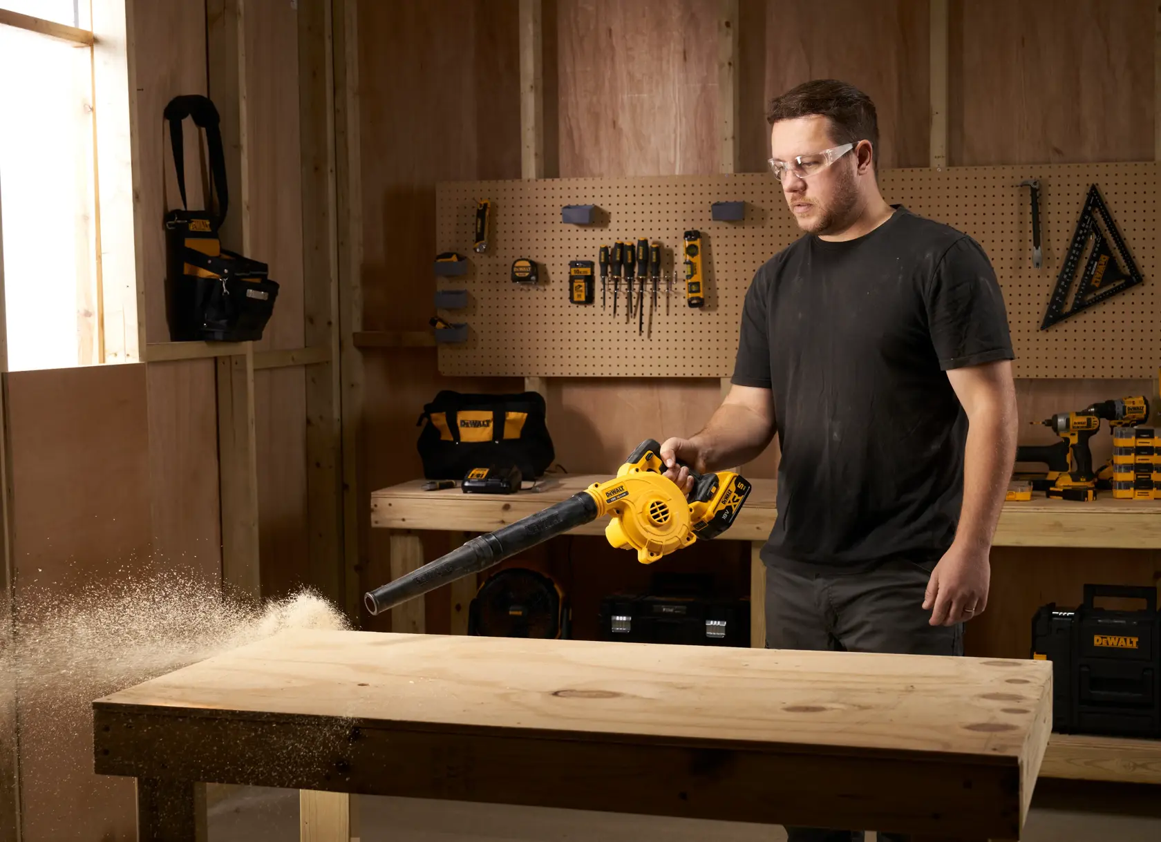 Persona limpia aserrín de un banco de madera con un soplador inalámbrico DEWALT en un taller.