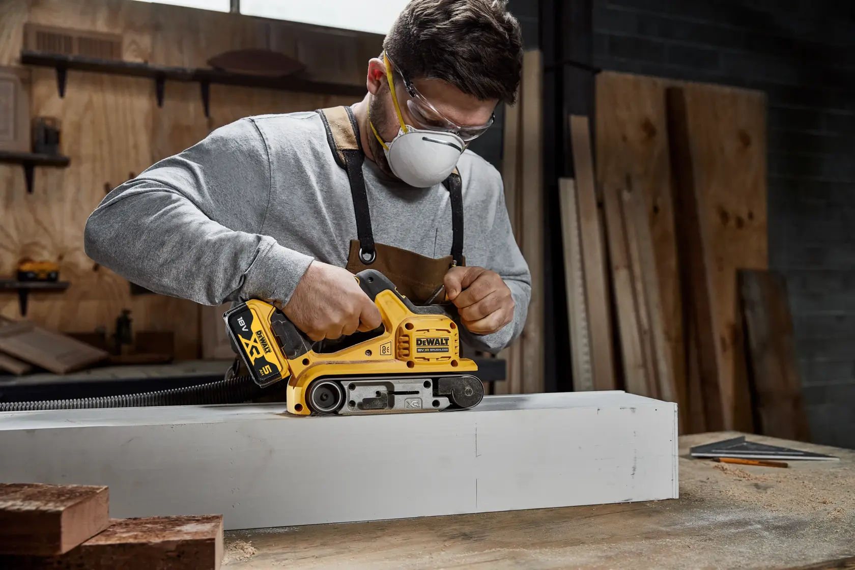 A person using a DEWALT cordless belt sander (SKU: DCW220P2) on a piece of wood in a workshop. The person is wearing safety goggles and an apron, and their face is blurred.
