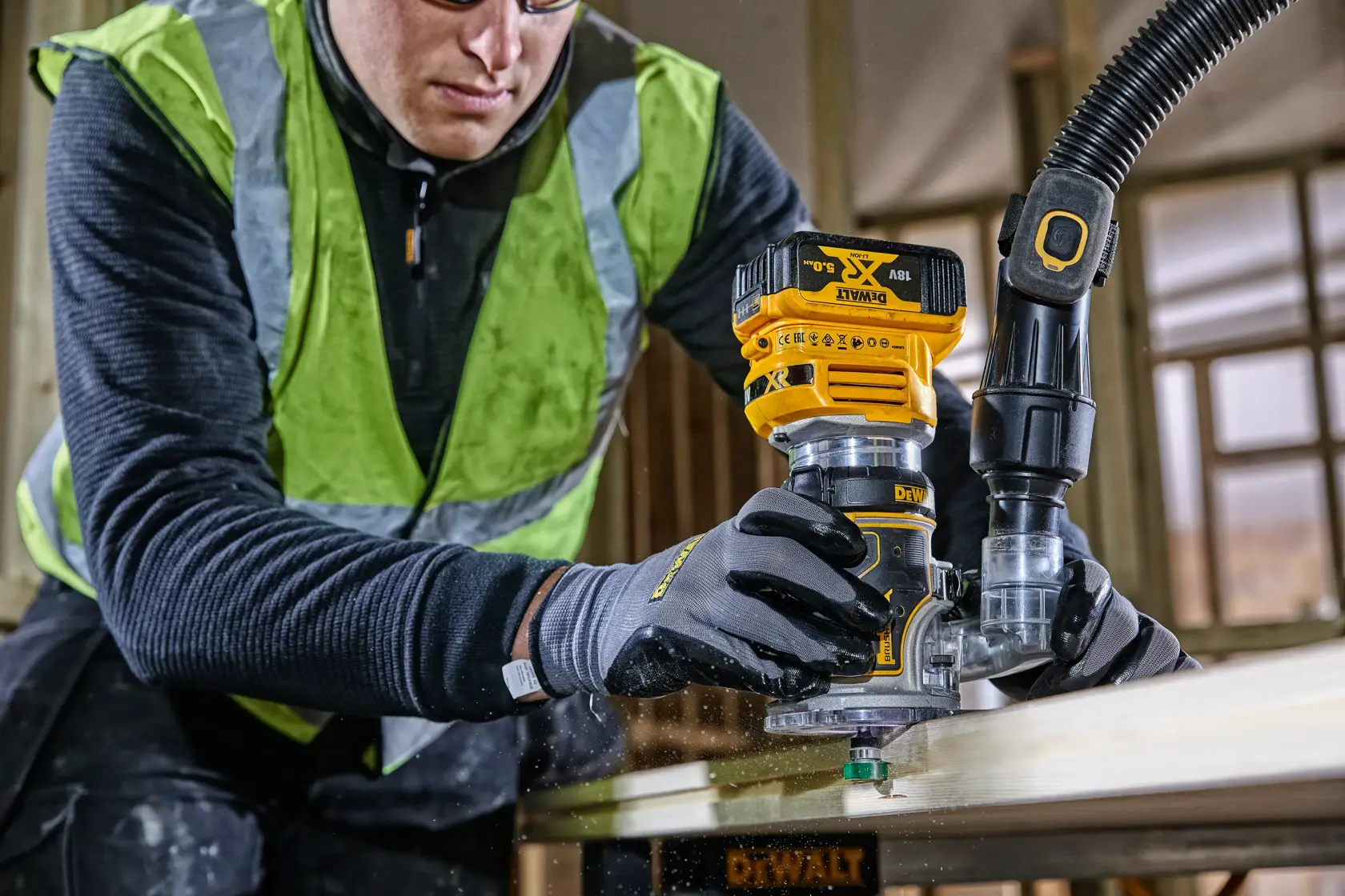 Ouvrier utilisant une défonceuse DEWALT DCW604 sur du bois, avec gants et gilet de sécurité.
