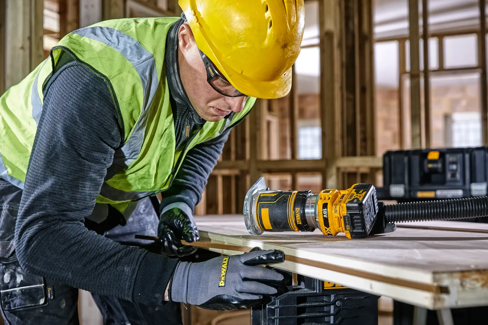Ouvrier utilise la défonceuse DEWALT DCW604 sur du bois, équipé de gants, casque et gilet de sécurité.