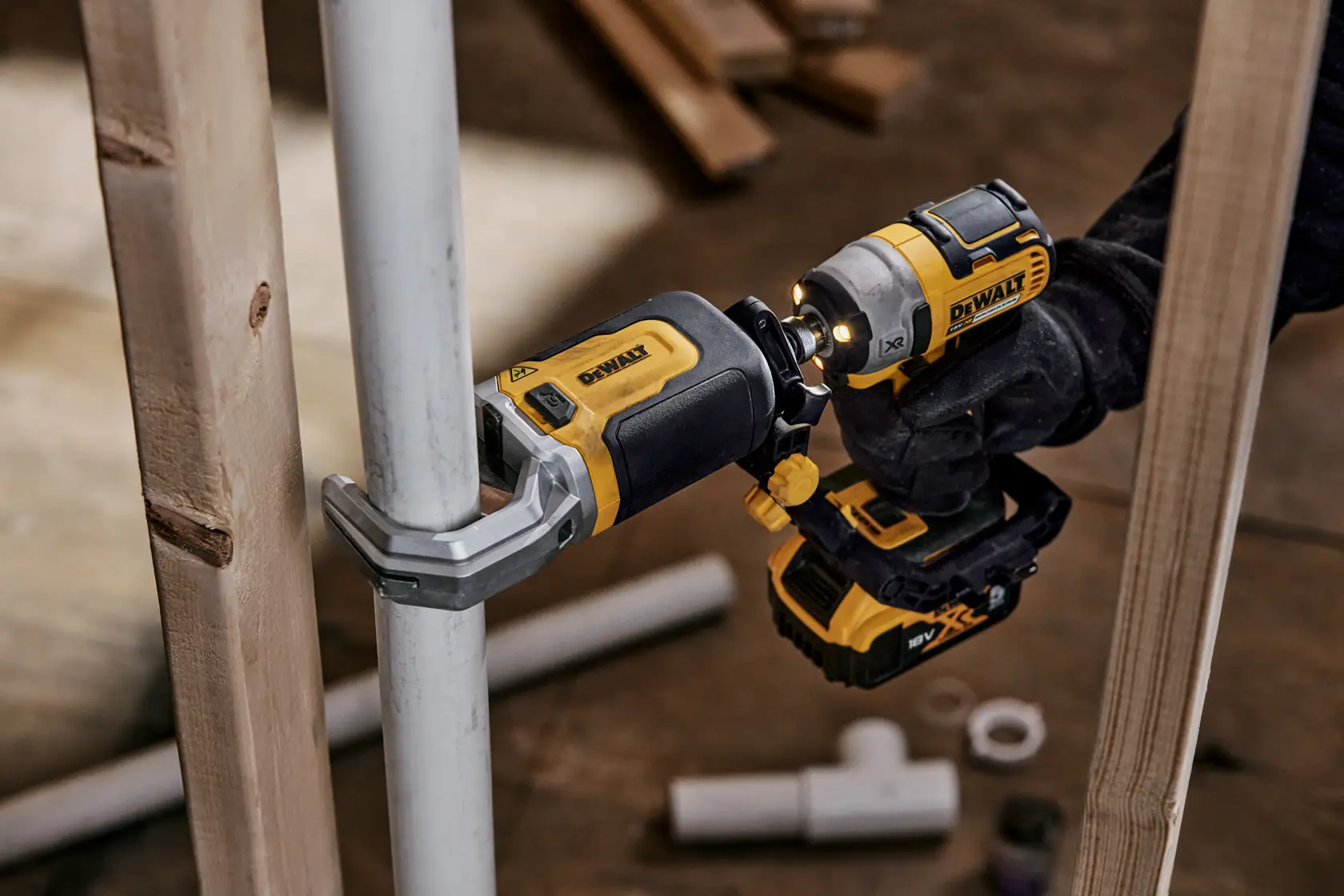 Una herramienta eléctrica DEWALT DT20560 usada para fijar una abrazadera a una tubería metálica, sostenida por una persona con guante. La herramienta es amarilla y negra, y en el fondo se ven otras tuberías y tablas de madera.