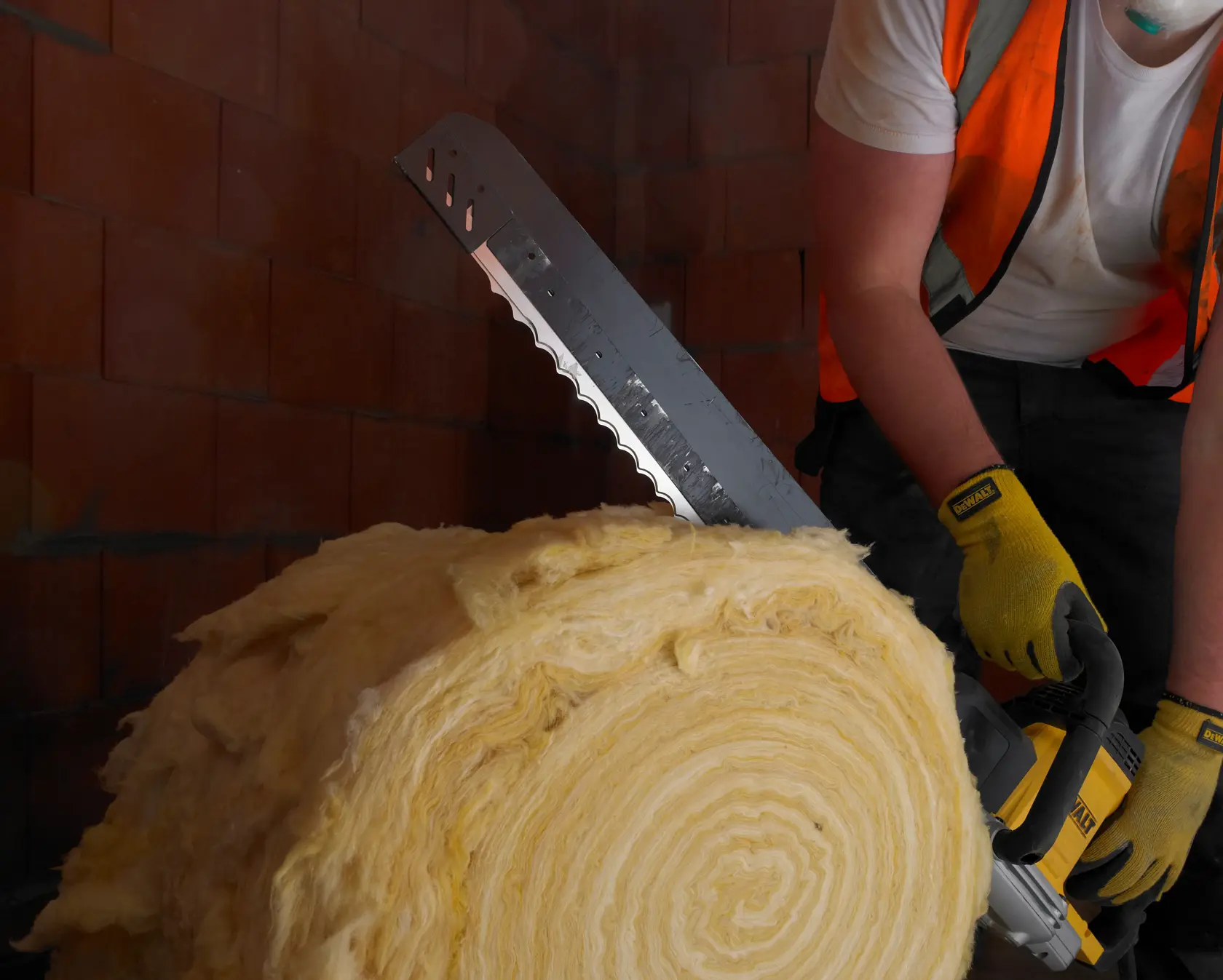 Person schneidet Isolierrolle mit DEWALT Säge, trägt Handschuhe und Weste, im Bereich mit Ziegelwand.