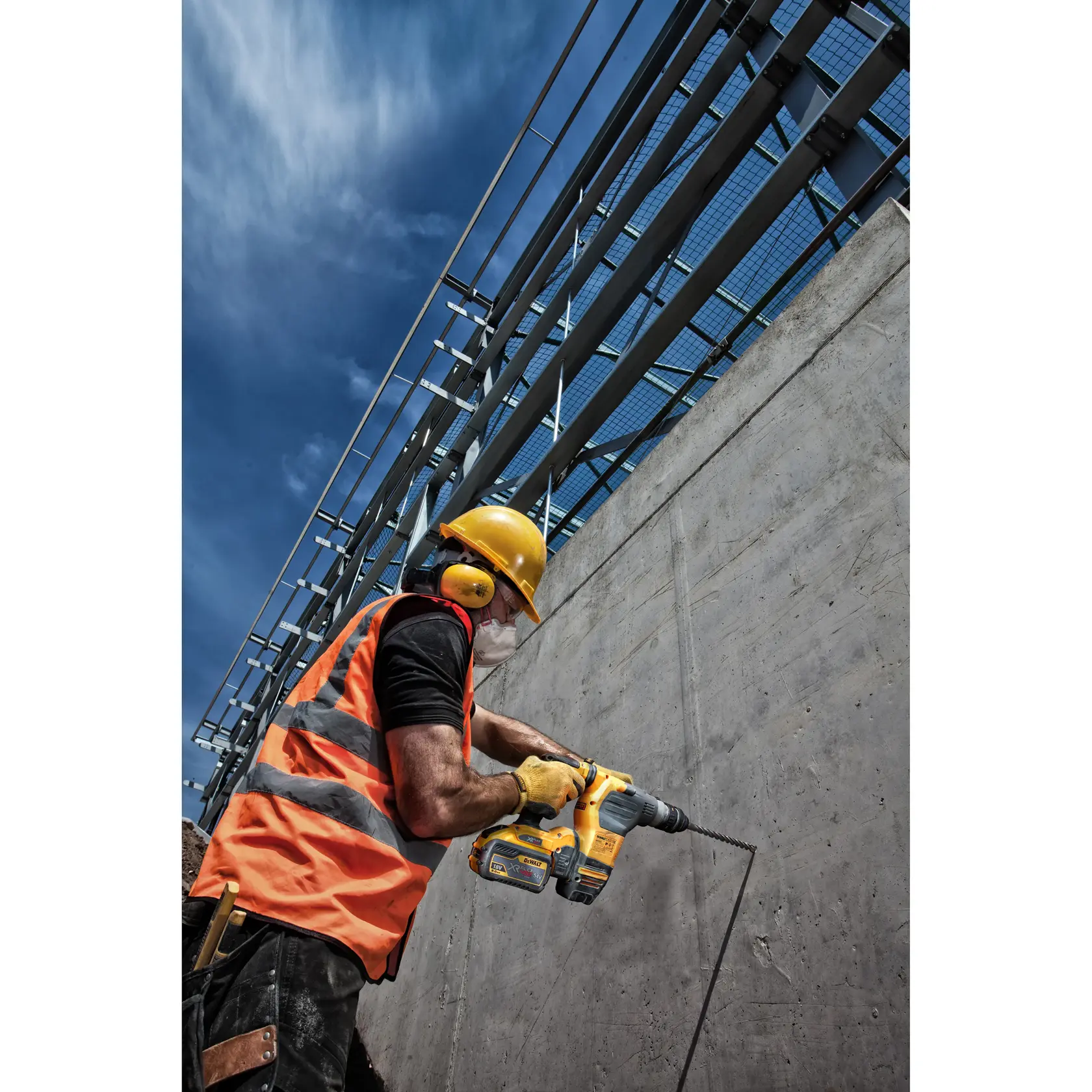 Trabajador usa taladro DEWALT en pared de concreto, con equipo de seguridad y chaleco naranja en obra.