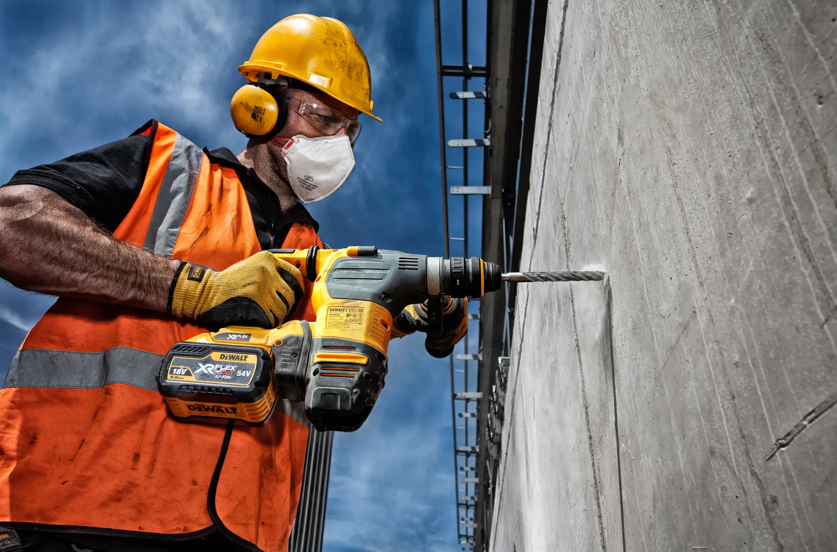 Worker drills concrete wall with DEWALT rotary hammer, wearing safety gear and orange vest outdoors.