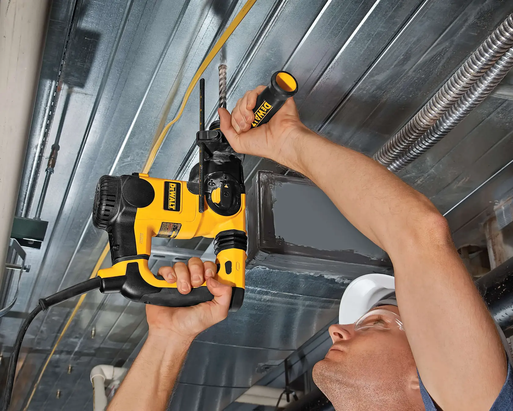 Photo of DEWALT DT9556 rotary hammer drill used overhead on metal ceiling, worker in hard hat visible.