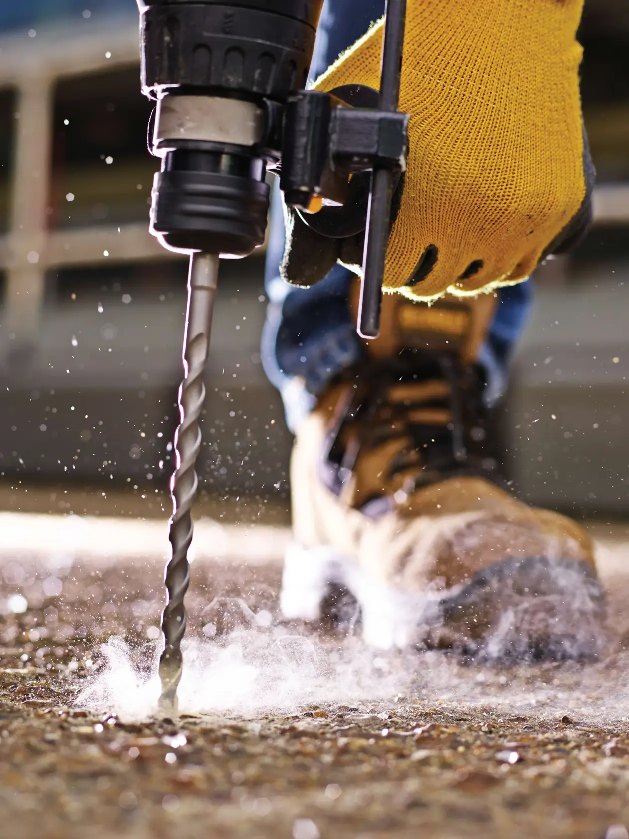 Close-up of a gloved hand using a drill bit on a concrete floor, with dust and safety boots visible.